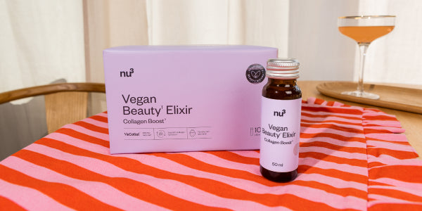 Emballage et flacon de Vegan Beauty Elixir sur une table avec une nappe rayée et un verre en arrière-plan