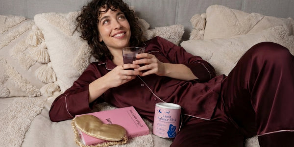 dunkelaarige Frau mit der Dose nu3 Luna Balance Elixir und einem Glas mit diesem Drink auf der Couch im Schlafanzug mit Buch und Schlafmaske